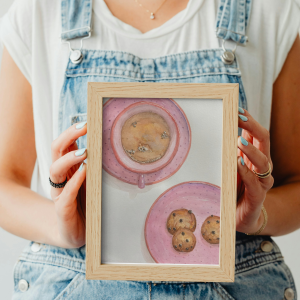 Art print akvarelne slike z roza skodelico kave in roza krožnikom s tremi piškoti. Slika je okvirju, ki ga v rokah drži ženska.